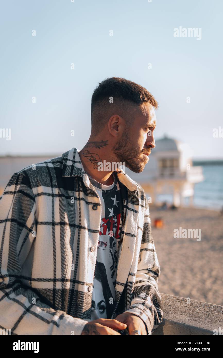 A portrait of a Caucasian young man in a plaid jacket and earring sitting with blur beach Stock Photo