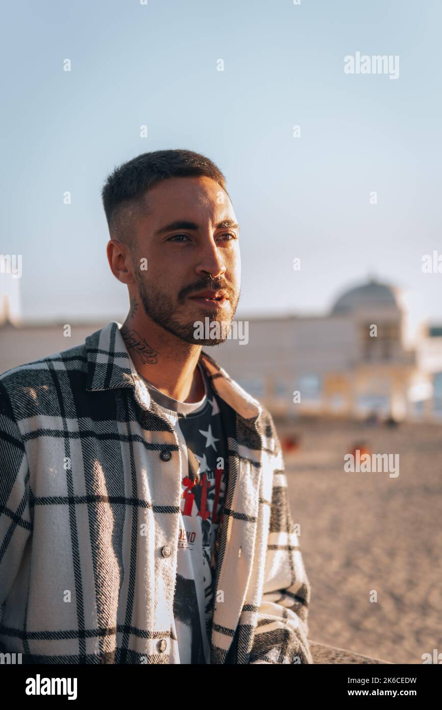 A portrait of a Caucasian young man in a plaid jacket with tattoo and earring sitting with blur beach Stock Photo