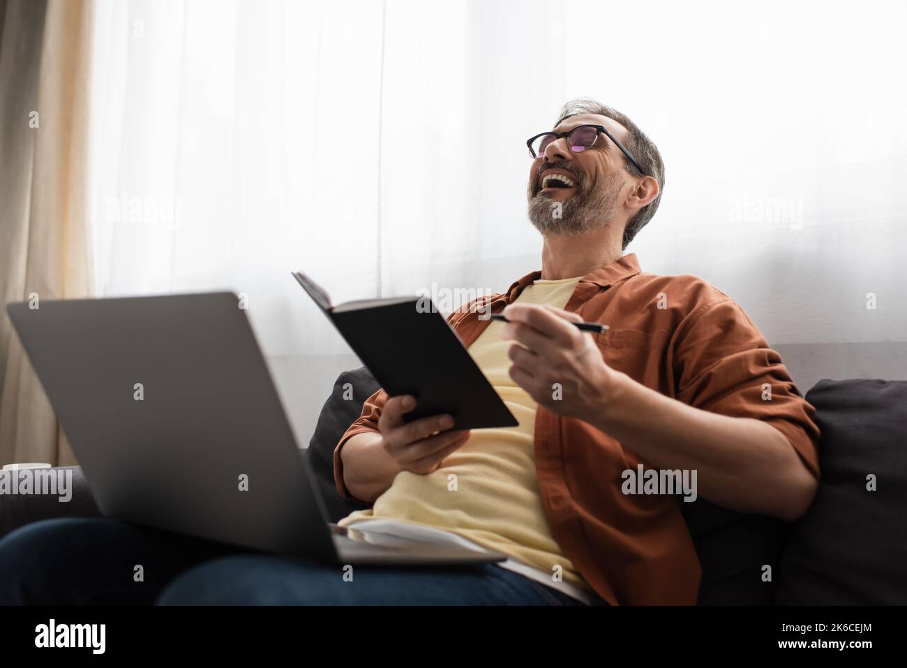 excited man in eyeglasses holding pen and notebook while laughing on couch near laptop,stock image Stock Photo