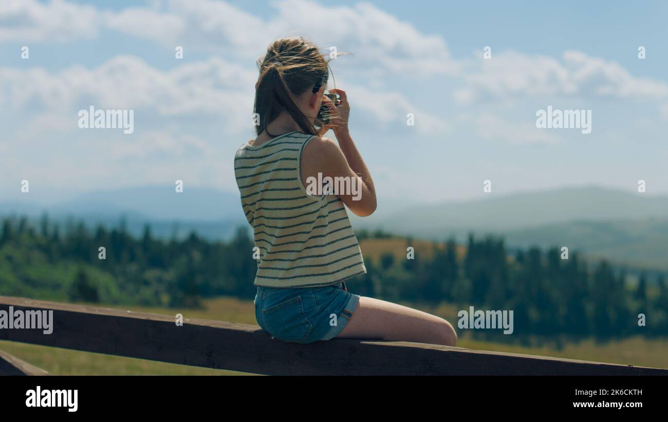 Young photographer shooting landscape on beautifull green hillway using digital camera, sitting on wooden fence, taking pictures of nature. Girl filling the portfolio with amazing photos. Stock Photo