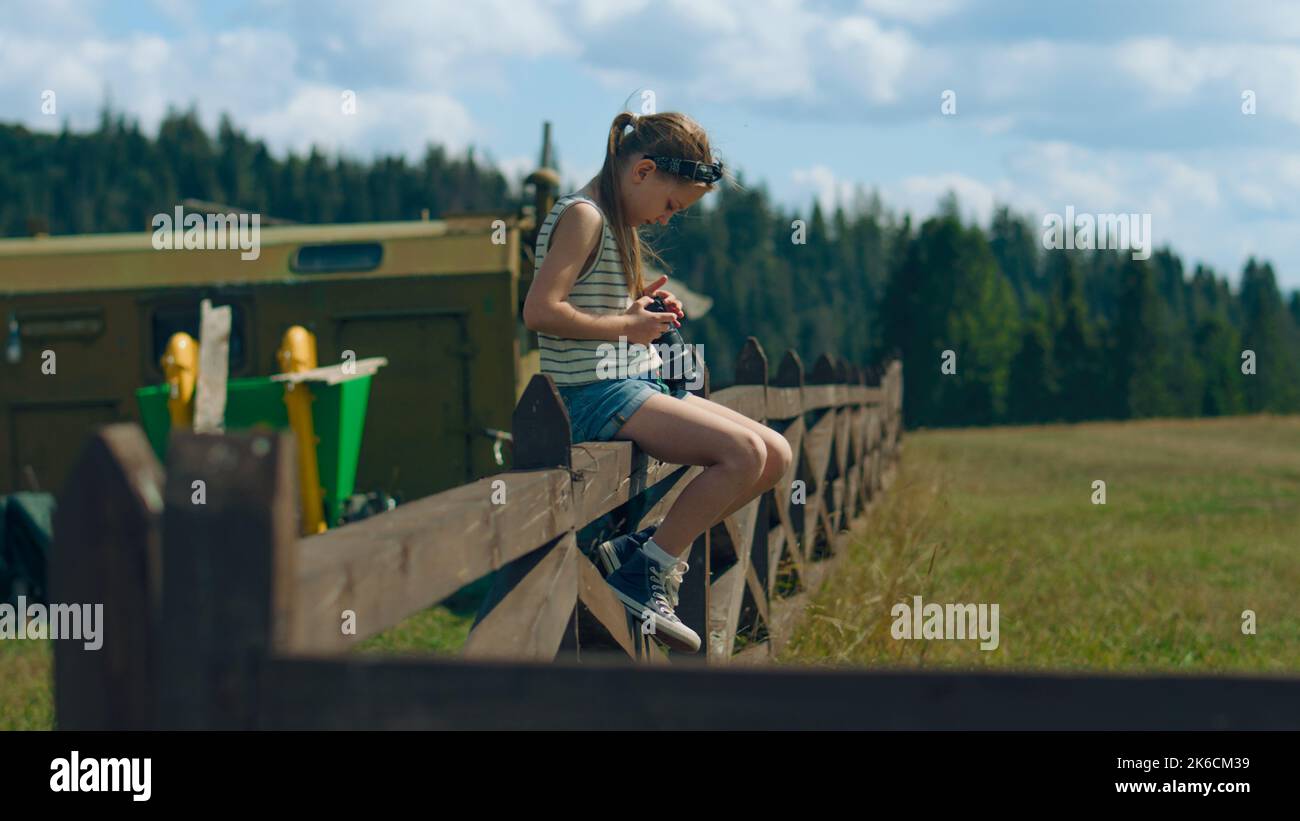 Young photographer wiping digital camera lens by t-shirt, sitting on wooden fence. Girl shooting beautiful nature landscape, concentrating on process, spending leisure time outdoor. Slow motion. Stock Photo