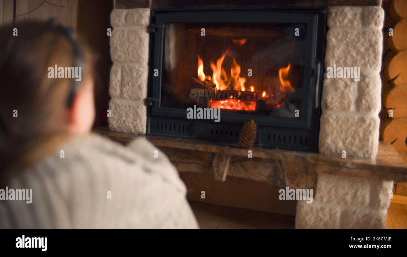 Young girl sitting by the fireplace on mild carpet, watching on fire and warming by coverlet in cold days, relaxing and spending leisure time at home. Back view. Stock Photo