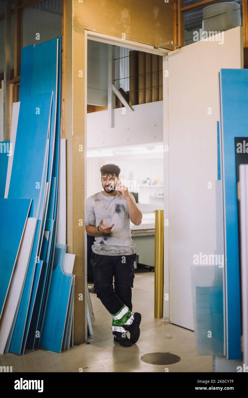 Full length of young male carpenter talking on smart phone while standing at doorway of workshop Stock Photo
