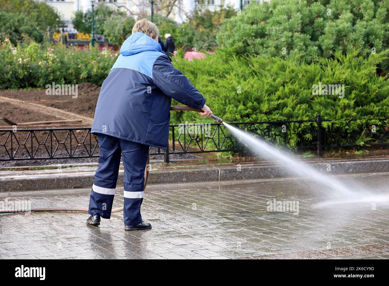 Woman worker in blue uniform watering the sidewalk with a hose. Street cleaning and disinfection in autumn city park Stock Photo
