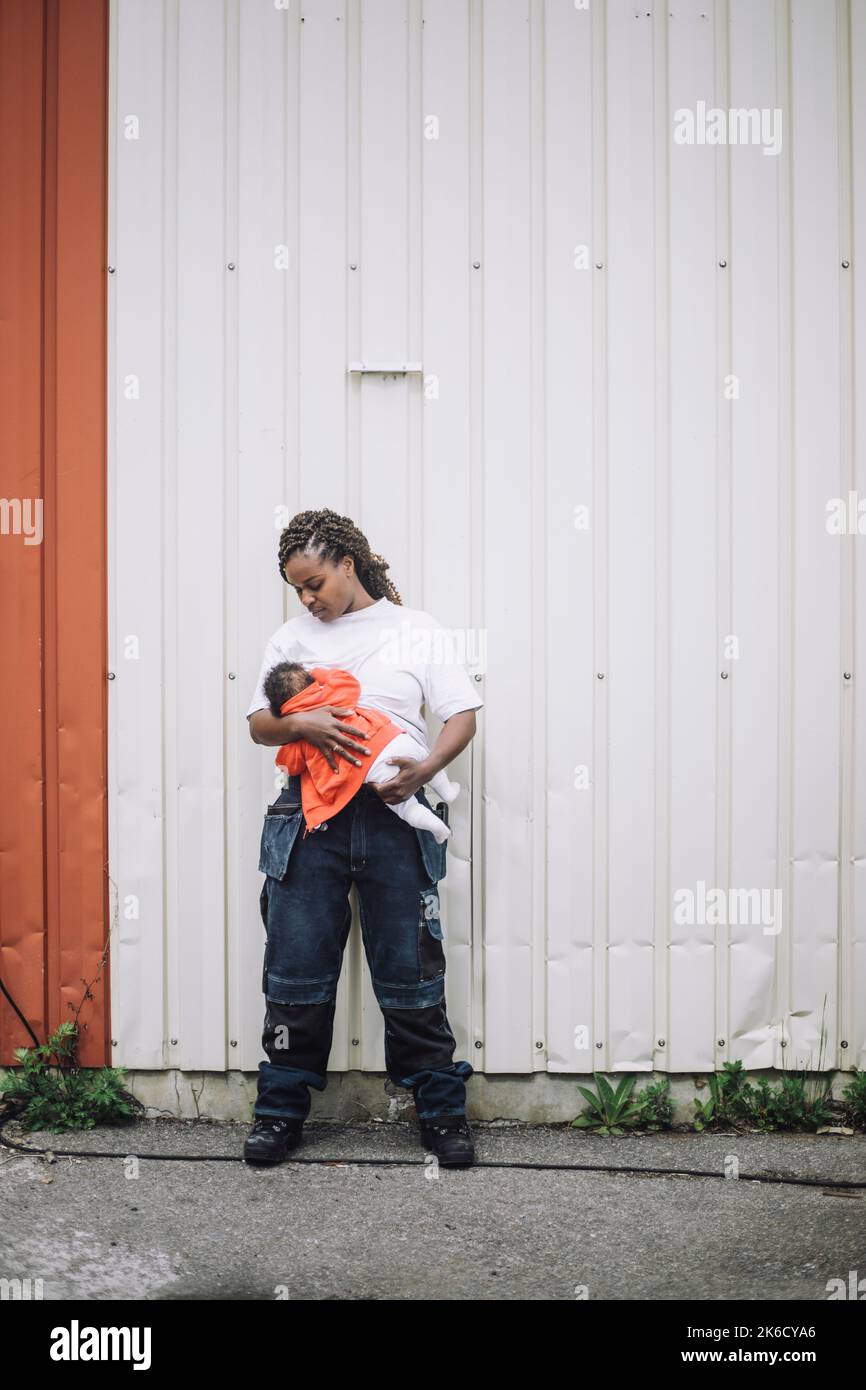 Full length of female construction worker breastfeeding daughter against metal wall Stock Photo