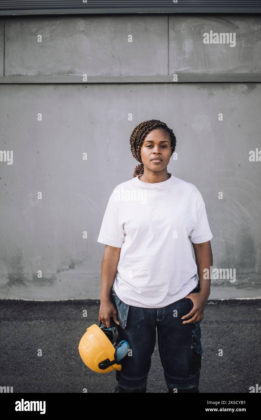 Female construction worker standing with hardhat against wall Stock Photo