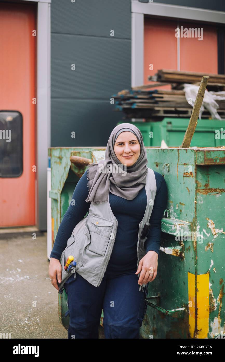 Smiling female construction worker wearing headscarf leaning on metal container Stock Photo