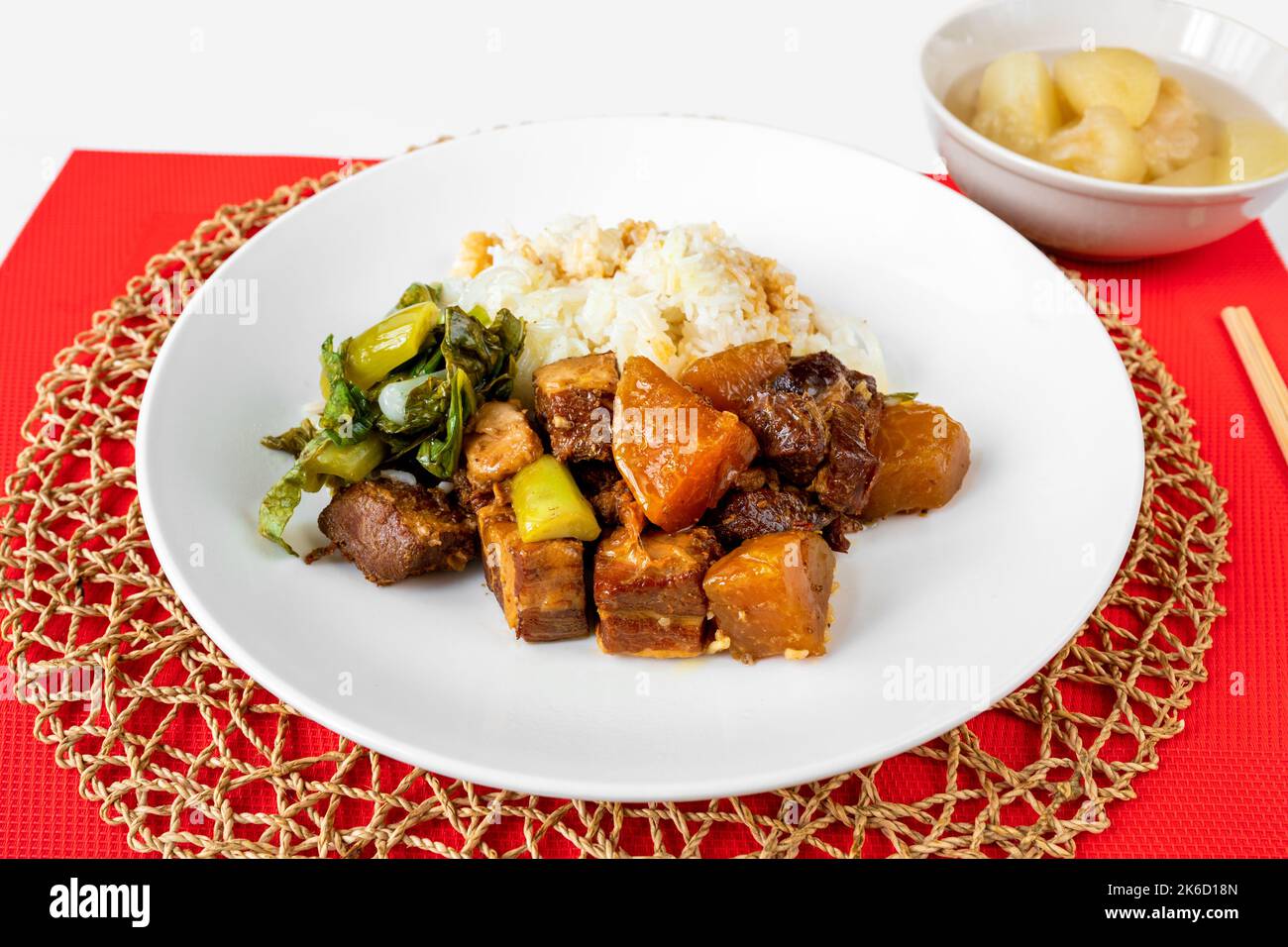 Caramelized cube of roasted pork belly, stewed cabbage and kohlrabi, rice on plate, chopstick on red table-cloth and circle bamboo pad, compote. Stock Photo