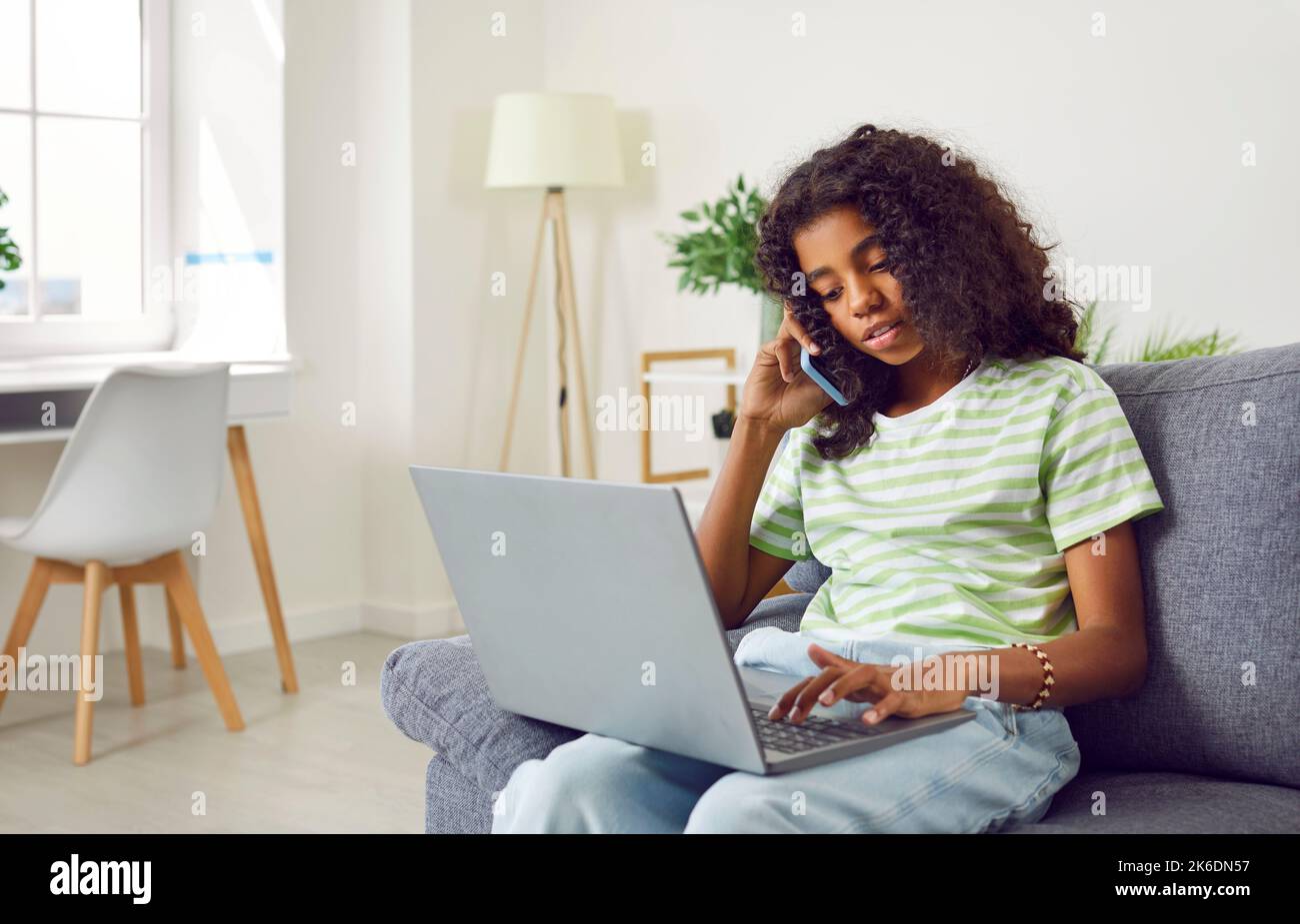 African america teen girl is calling mobile phone and using laptop sitting on couch at home. Stock Photo