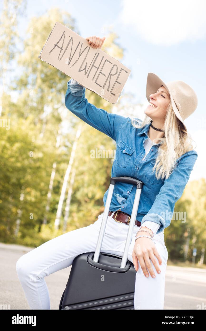 Young smiling blond woman raising looking at cardboard with inscription anywhere, sitting on black suitcase near forest. Stock Photo