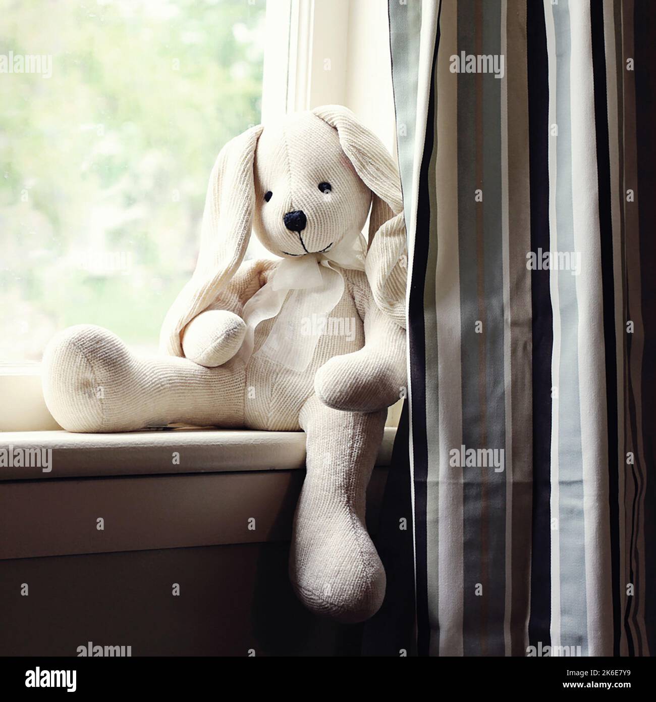 A soft white and cream stuffed animal rabbit keeps watch sitting in the window with outside light Stock Photo
