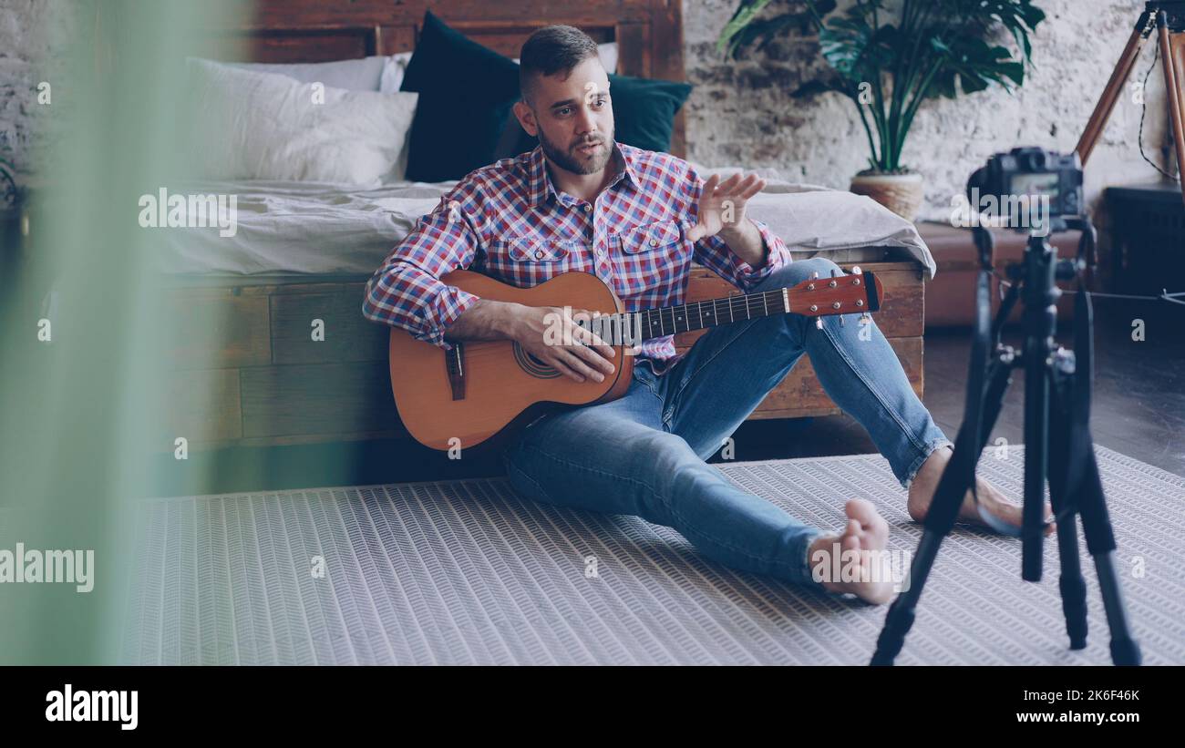 Handsome man blogger and guitarist is recording video lesson about guitar in bedroom using camera. Bearded guy is greeting his followers, talking and showing guitar parts. Stock Photo