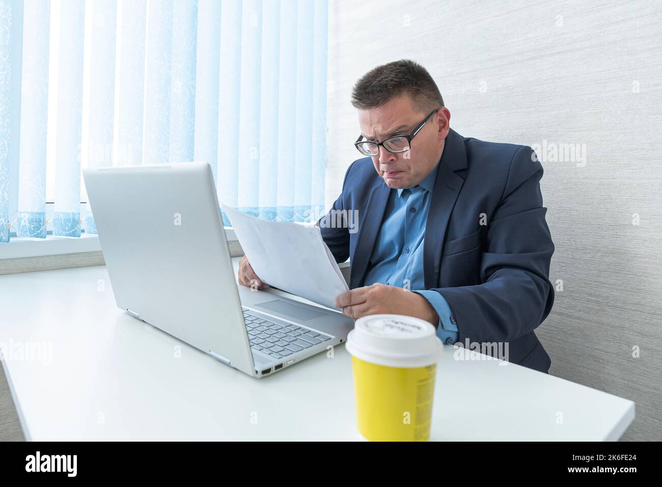A male employee in a blue jacket works with documents and on a computer in a bright office. Surprised emotion on the face. Stock Photo