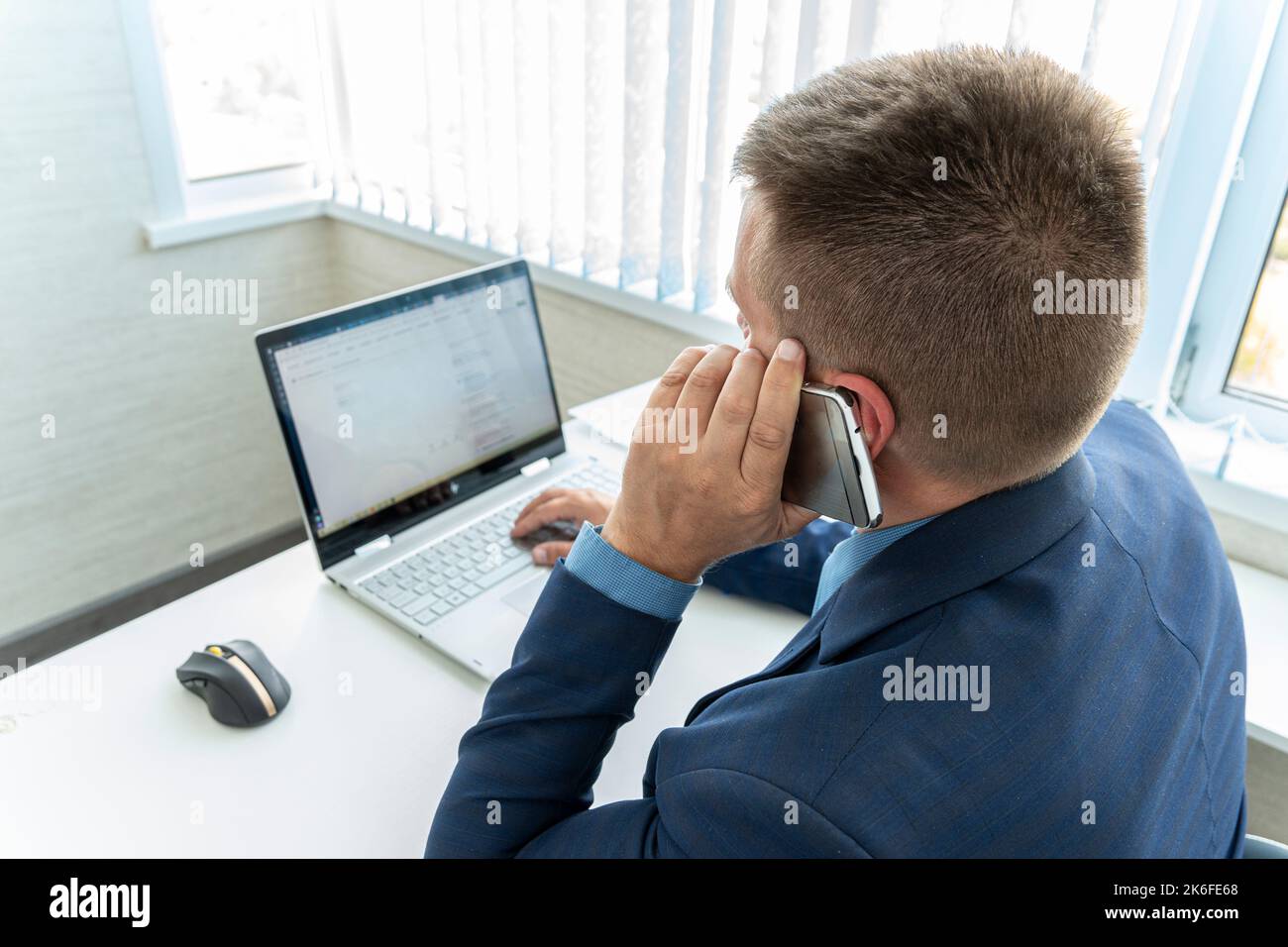 Concentrated businessman having a phone call and working on his laptop in his office. A manager in a bright office dressed in a blue business suit tal Stock Photo