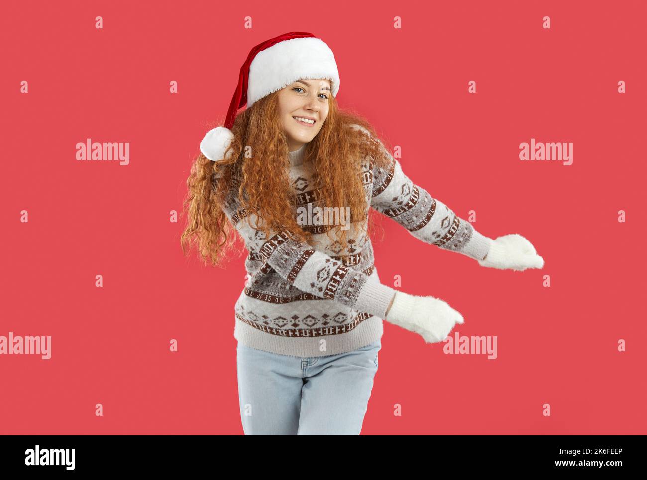 Cheerful smiling woman in sweater, Christmas hat and mittens dancing on red background Stock Photo