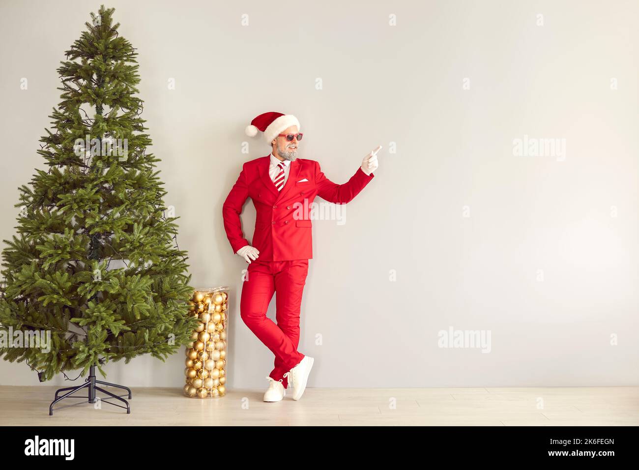 Funny man in red suit standing next to Christmas tree and pointing at copy space wall Stock Photo