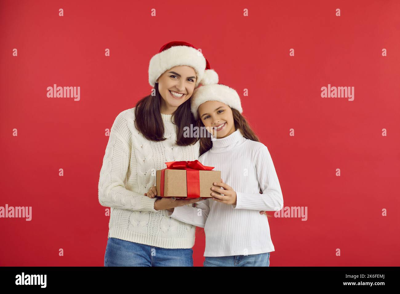 Happy young mom and teen daughter holding christmas present while standing on red background. Stock Photo