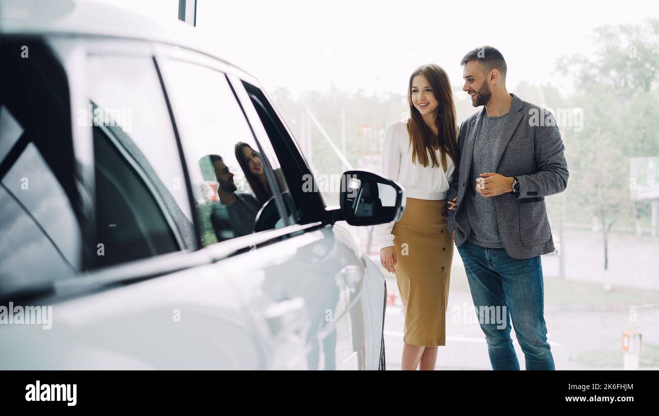 Attractive young couple confident bearded guy and his smiling pretty girlfriend are choosing new automobile together looking at luxury car in auto dealership and talking. Stock Photo