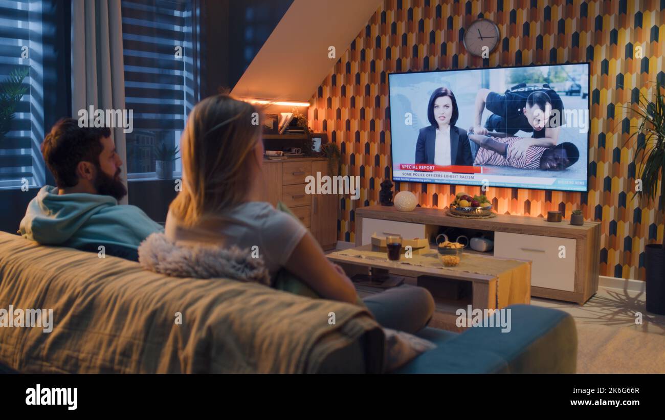Couple sitting on sofa in living room with snacks, discussing TV news special report about racism and discrimination in the police. Spending time in the evening at home together. TV live broadcasting Stock Photo