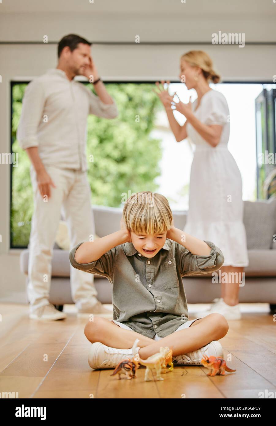 Young caucasian boy shouting, crying and covering his ears to block out the noise of his mother and father arguing in the background. Fighting impacts Stock Photo