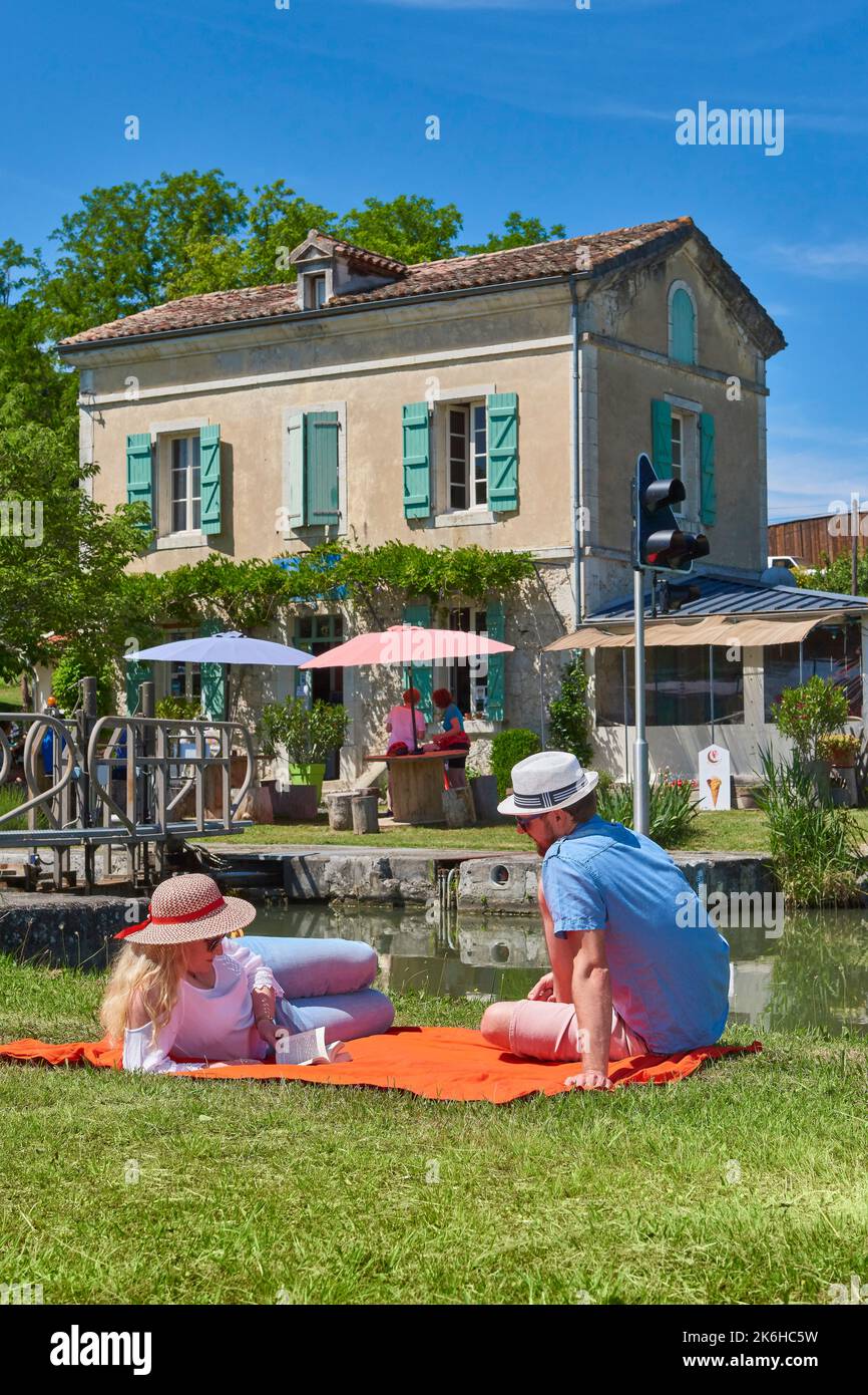 Couple relaxing and reading on the banks of the Canal du Midi in front of the lock keeper’s house converted into a restaurant and bed and breakfast ca Stock Photo