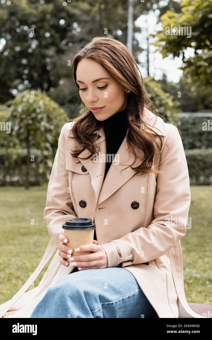 young woman in beige trench coat sitting on bench with paper cup,stock ...