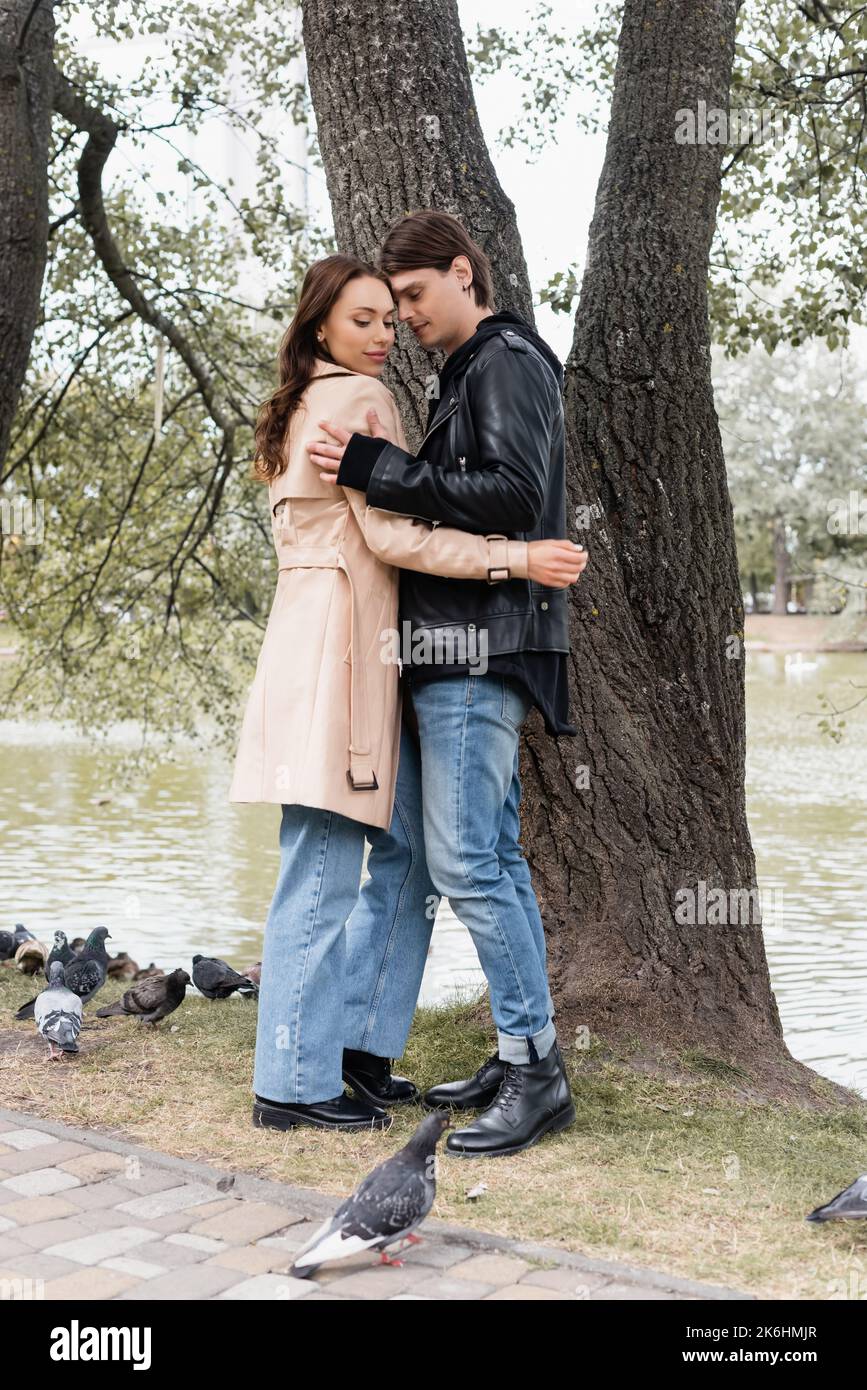 full length of stylish man and pretty woman in trench coat hugging near ...
