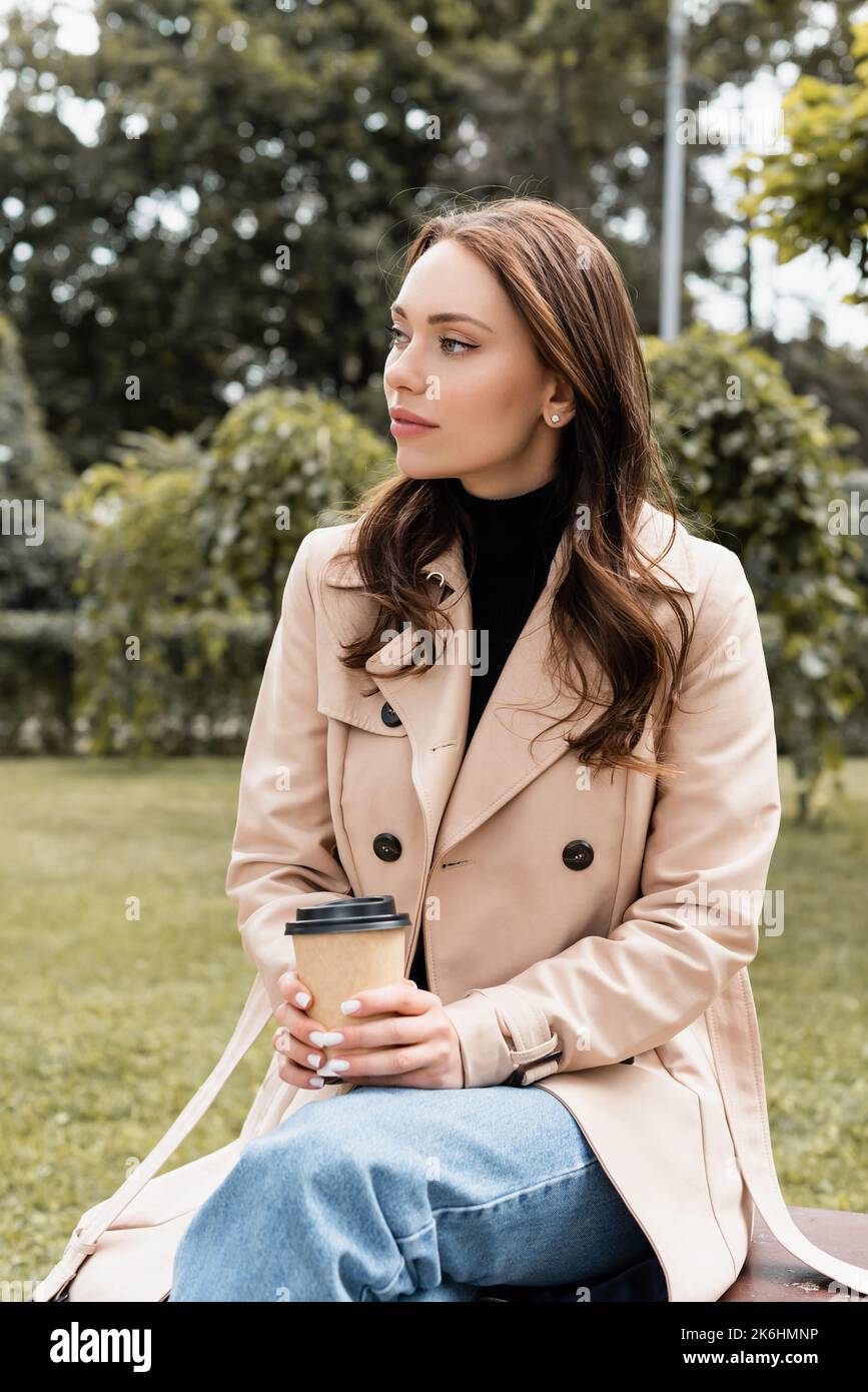 pretty young woman in beige trench coat sitting on bench with paper cup ...