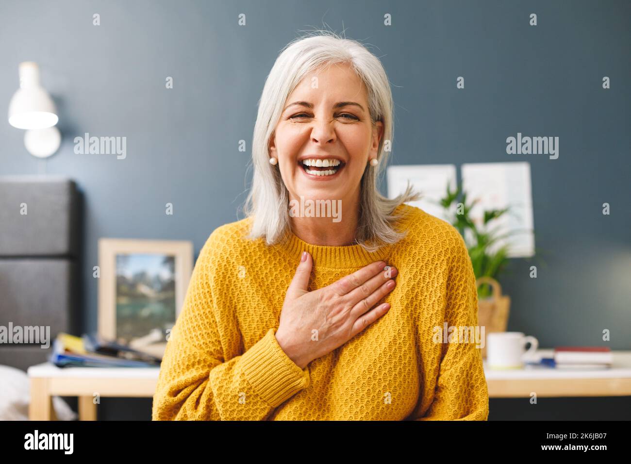 Happy caucasian senior woman making video call at home smiling to camera and touching chest Stock Photo