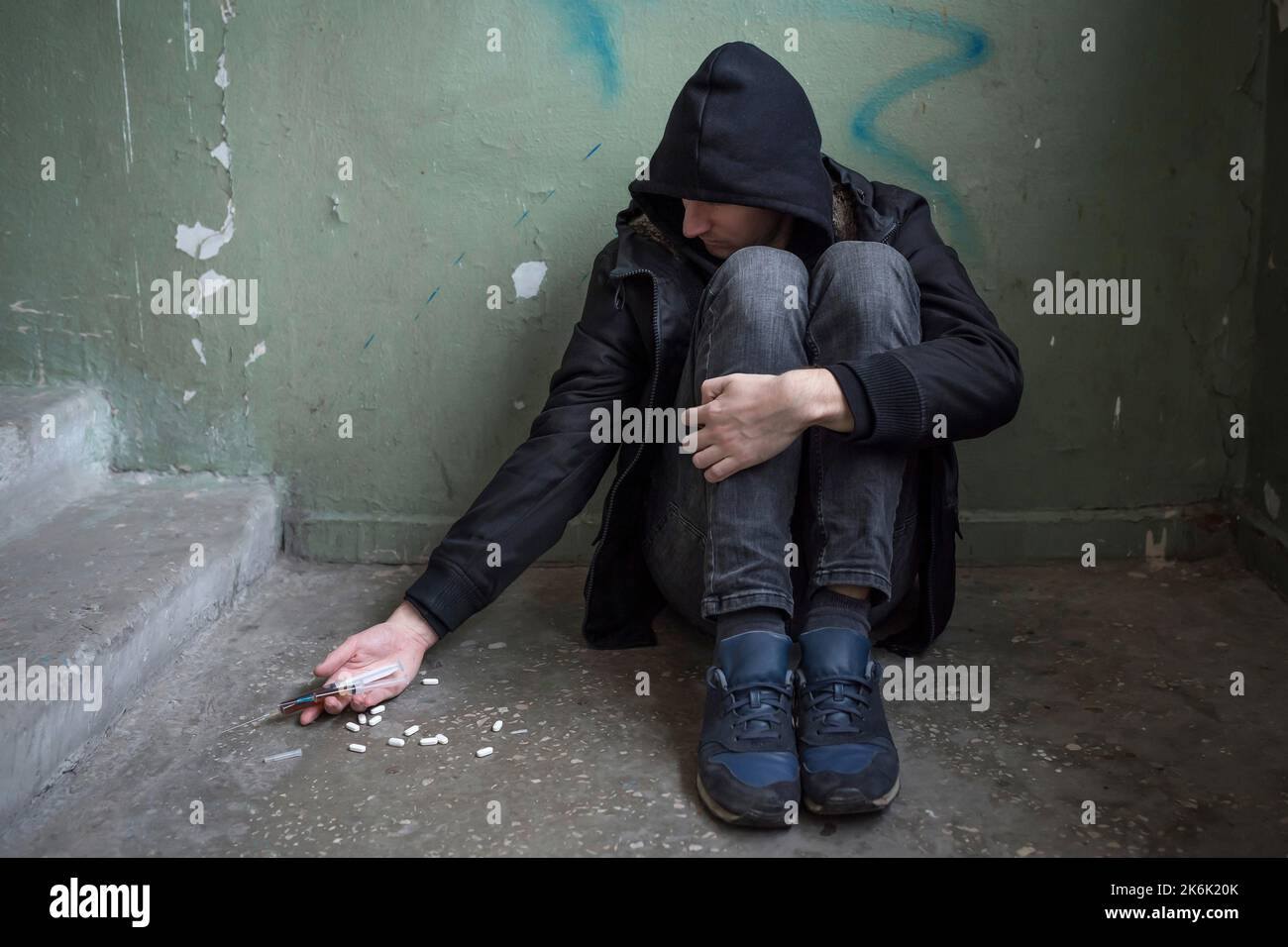 A male drug addict with a syringe is using drugs sitting on the floor. International Day Against Drug Abuse. Addiction concept. Stock Photo