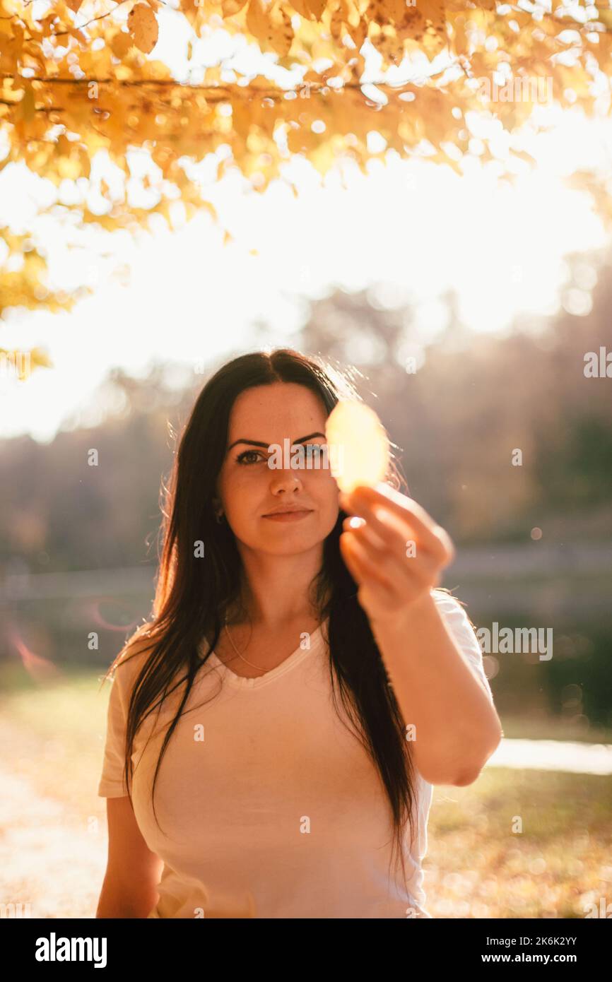 Beautiful young woman holding yellow leaf in front of her face while ...