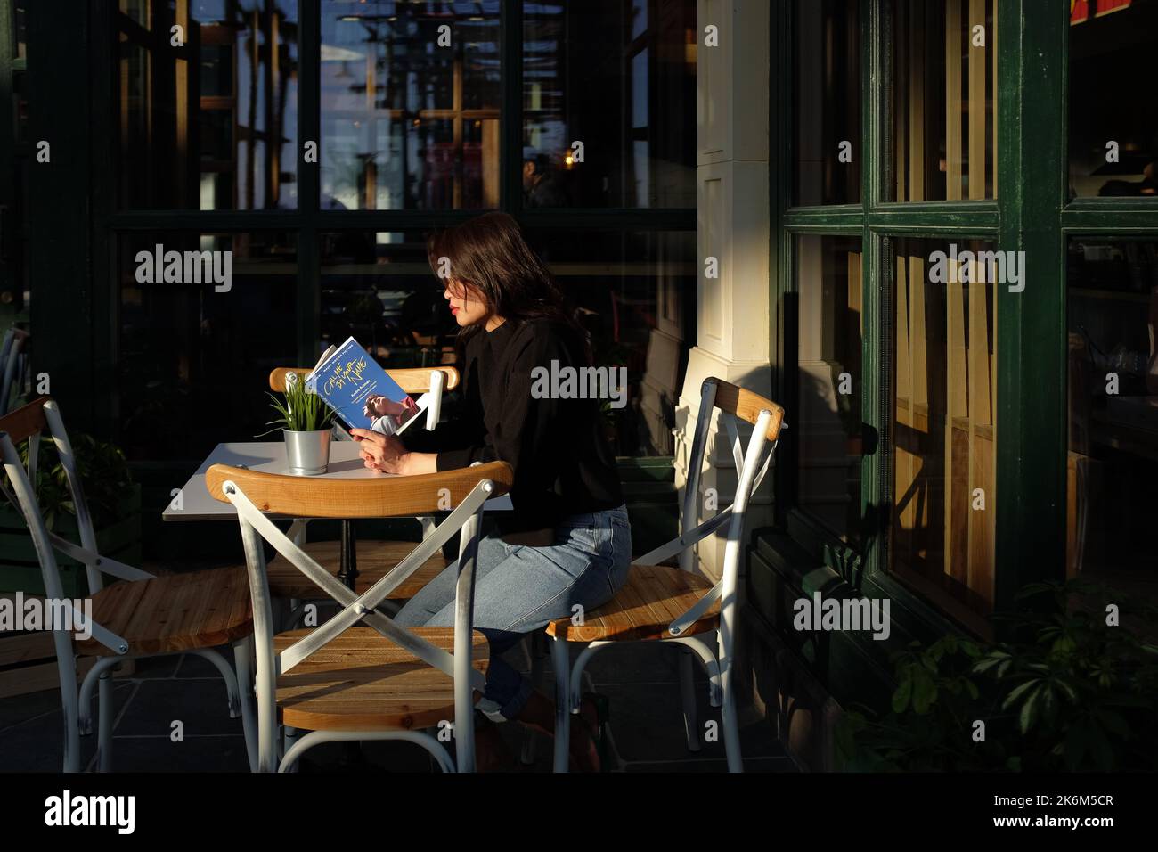 Asian woman wearing dark sweater and light jeans sits on a chair outside a cafe. Female is reading a book, outdoors, on a beautiful summer day. Stock Photo