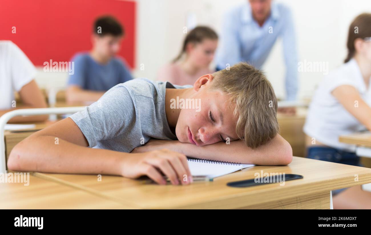 Bored teenage boy sleeping at desk in classroom during lesson Stock Photo