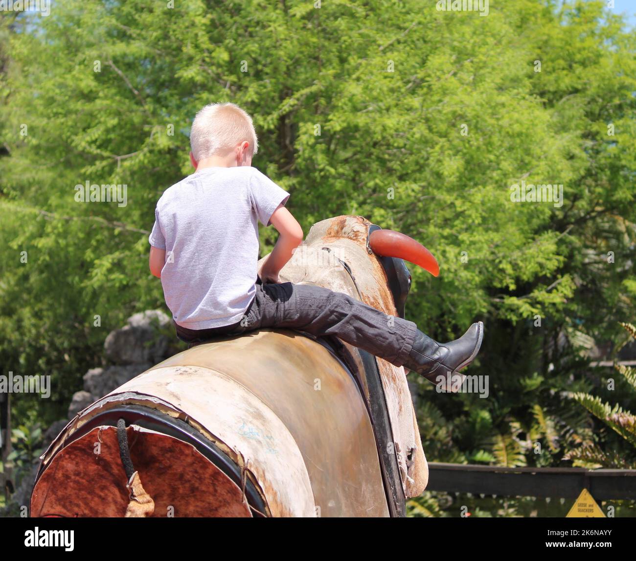 A young boy, wearing cowboy boots, riding on the back of a mechanical bull. Stock Photo