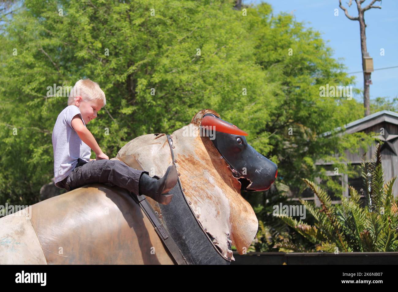 A young boy, wearing cowboy boots, riding on the back of a mechanical bull. Stock Photo