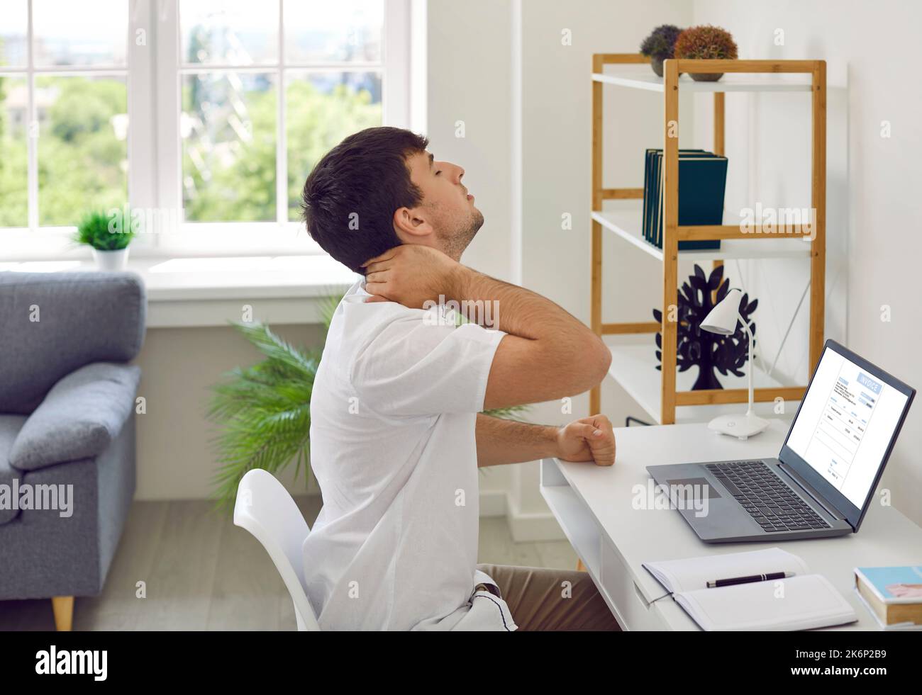 Tired, stressed man sitting at working desk with laptop and holding hand on his painful neck Stock Photo