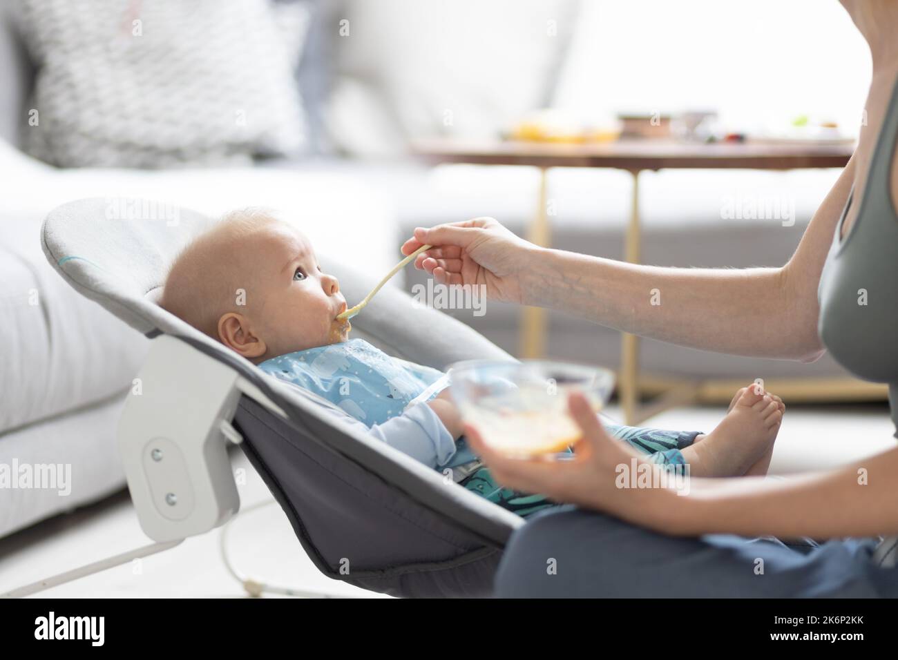 Mother spoon feeding her baby boy infant child in baby chair with fruit
