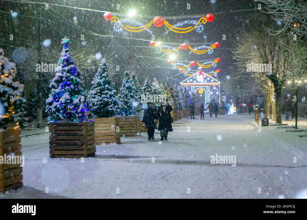 Snowy city street with Christmas trees and illumination decoration during snowfall on winter night. People walking down street. Abstract Christmas New Year and winter background. Stock Photo