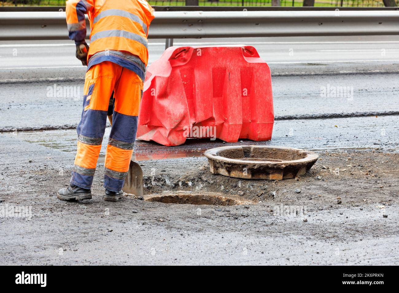A road worker in orange overalls clears the mouth of a sewer manhole on the carriageway with a shovel for its subsequent repair. Copy space. Stock Photo
