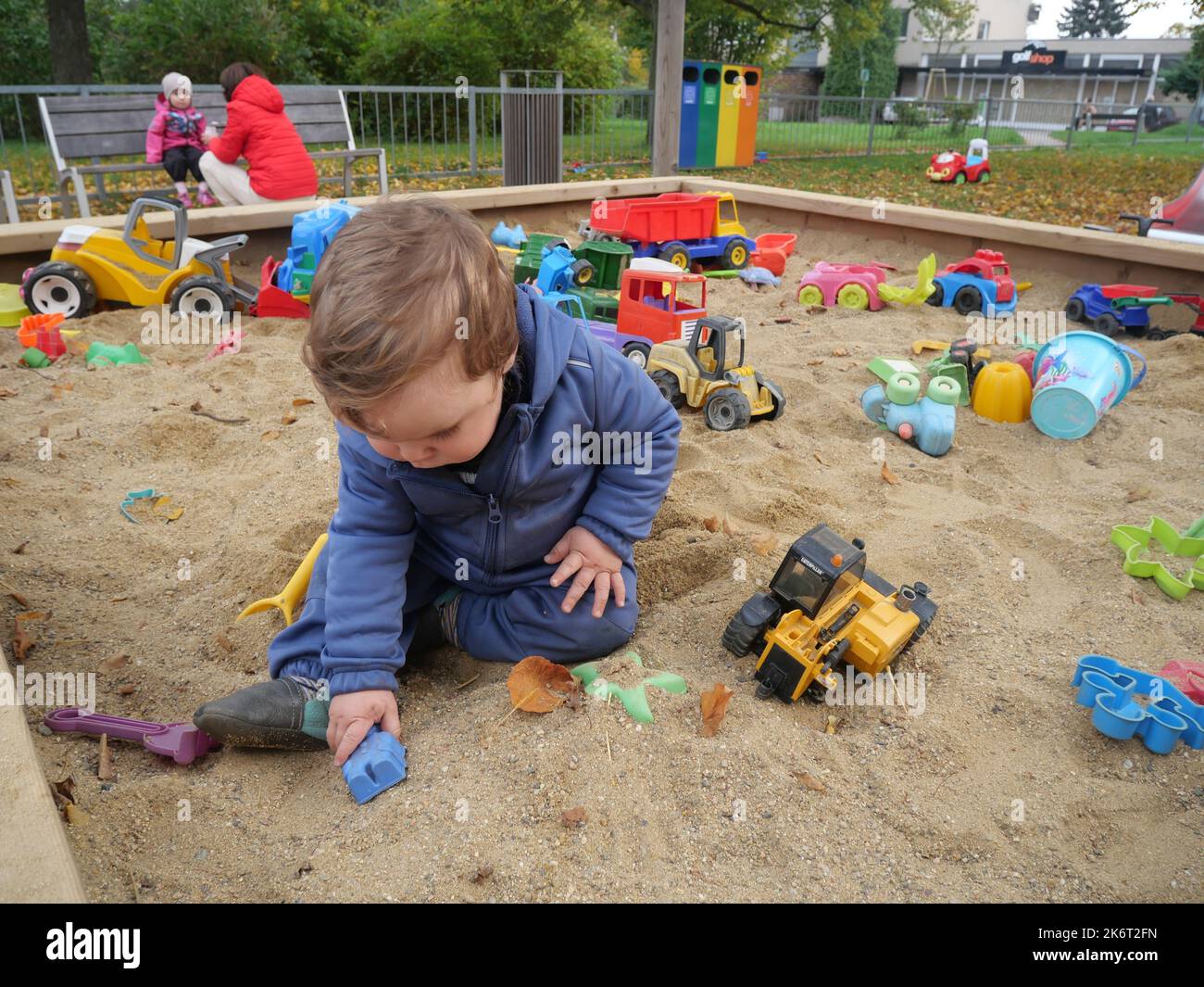Cute small boy toddler playing in the sand on a playground. Child sitting in a sandpit holding a plastic toy, with other toys in the background. Stock Photo