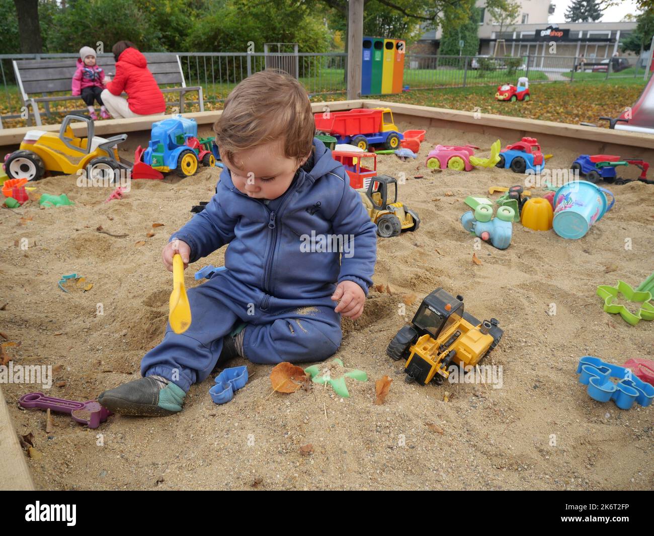 Cute small boy toddler playing in the sand on a playground. Child sitting in a sandpit holding a plastic toy, with other toys in the background. Stock Photo