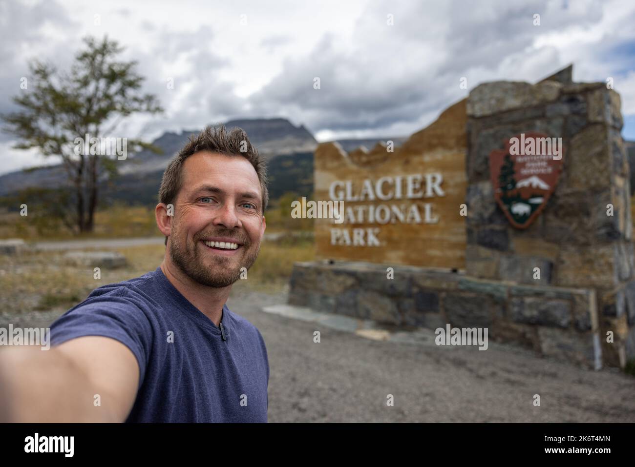 Person with a microphone clipped to his shirt taking a selfie in front of the east entrance sign for Glacier National Park Stock Photo