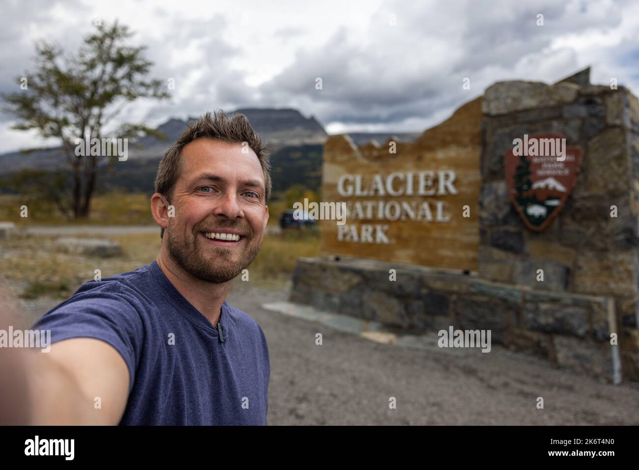 Person with a microphone clipped to his shirt taking a selfie in front of the east entrance sign for Glacier National Park Stock Photo