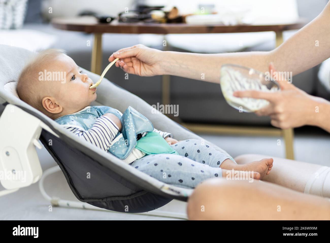 Mother spoon feeding her baby boy infant child in baby chair with fruit puree. Baby solid food introduction concept. Stock Photo