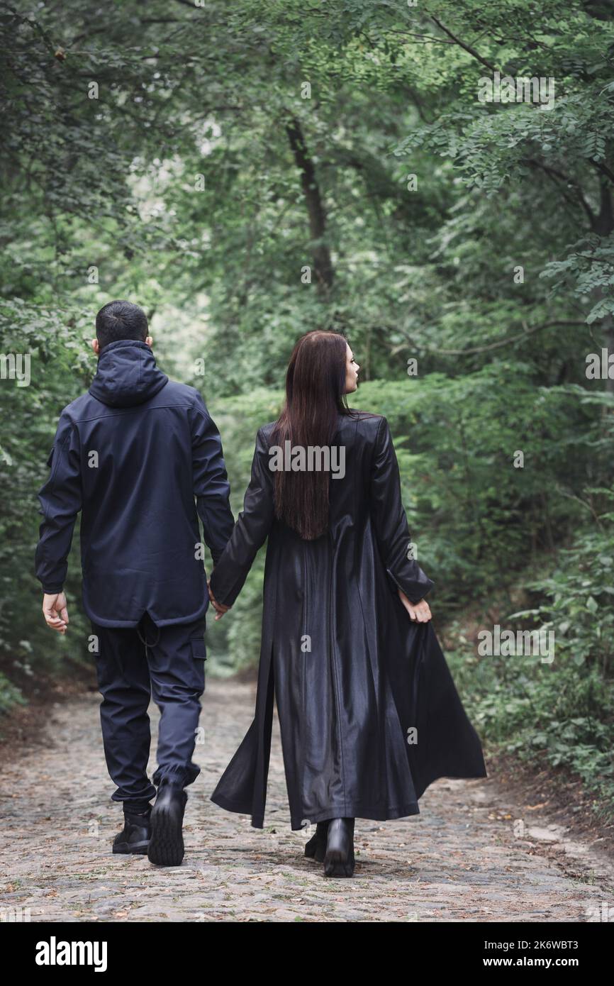 A guy and a girl in black brutal clothes are walking in a dark, foggy forest, back view Stock Photo