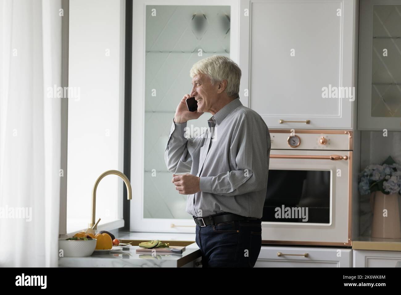 Older single man talking on smartphone standing in kitchen Stock Photo