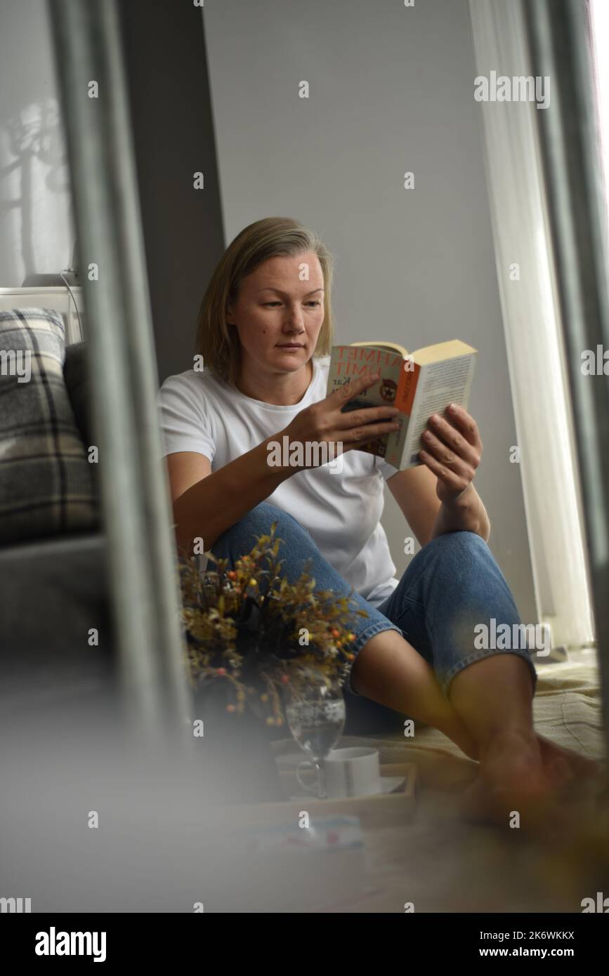 Beautiful woman sitting at apartment floor near mirror and feeling interested on literature book, beautiful woman holding poem recreating in home Stock Photo