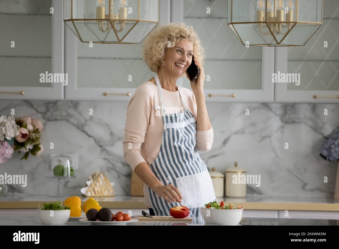 Older woman standing in kitchen talks on smart phone Stock Photo