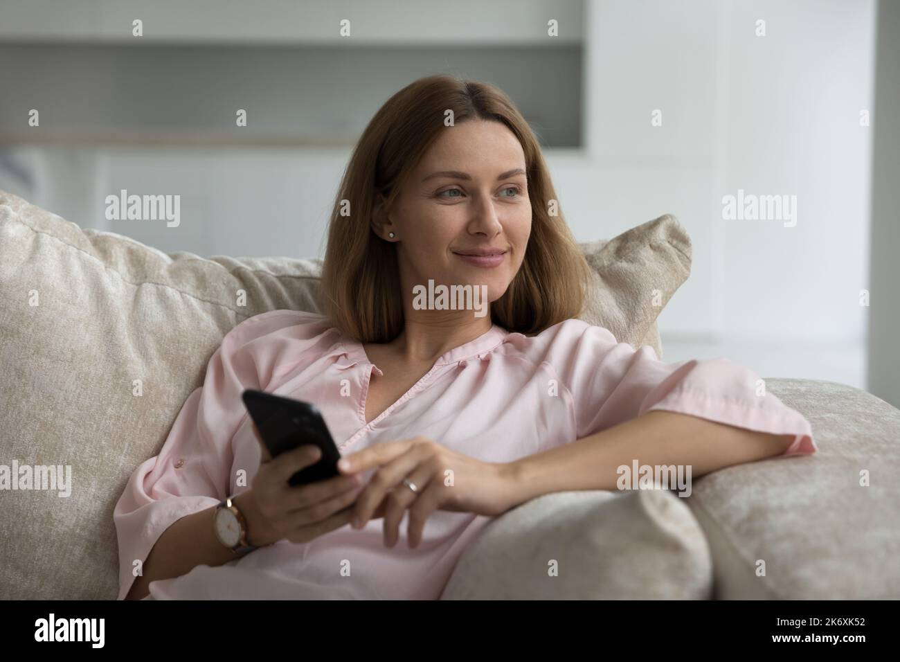 Woman looks aside sits on sofa with smartphone in hands Stock Photo