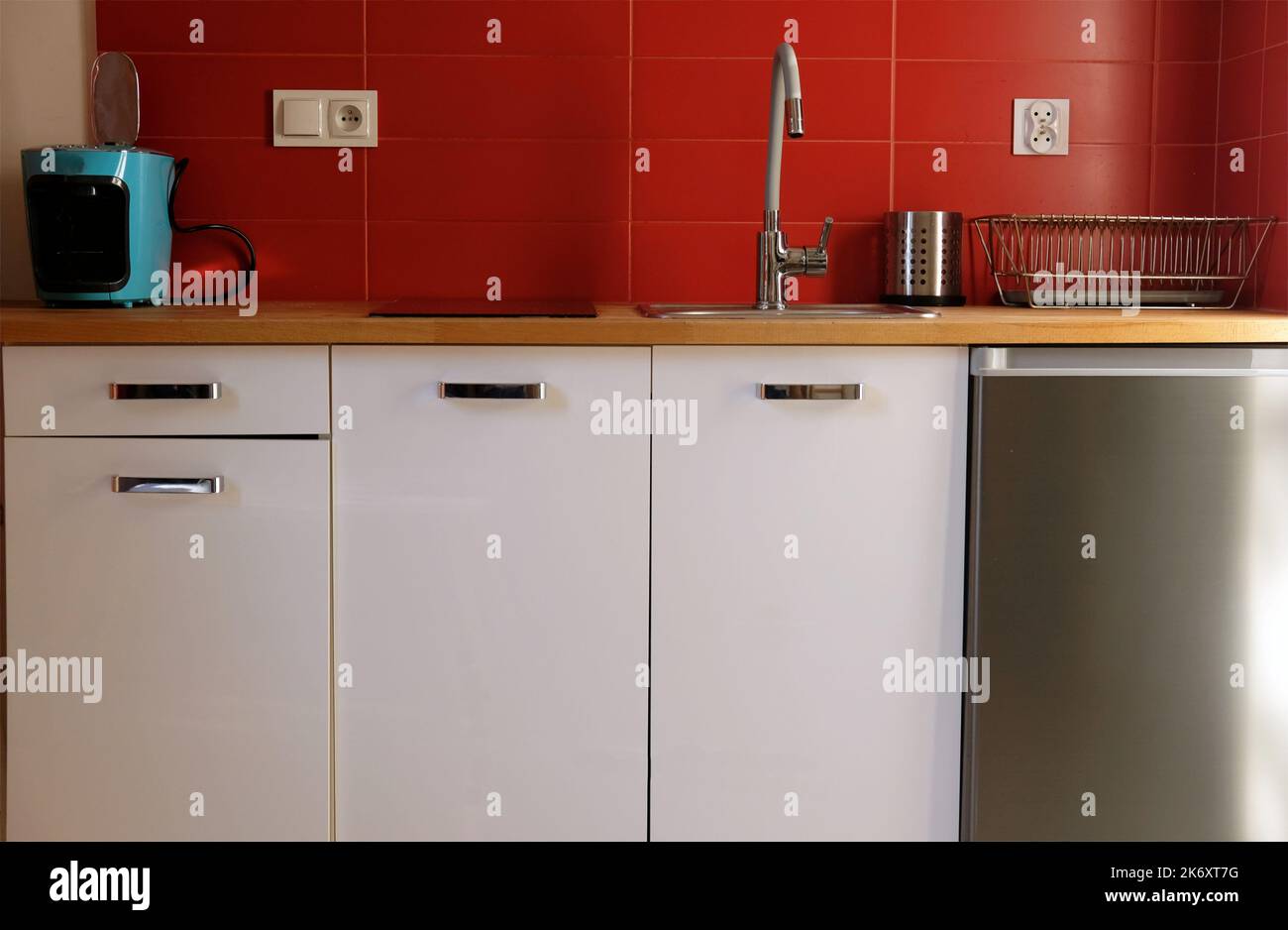 row of modern white kitchen cabinets with red tile wall, woodent worktop and stainless steel appliances fridge in a small apartment Stock Photo